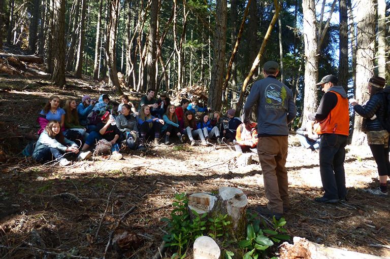 Three adults presenting to a group of young people seated on the ground in a forest